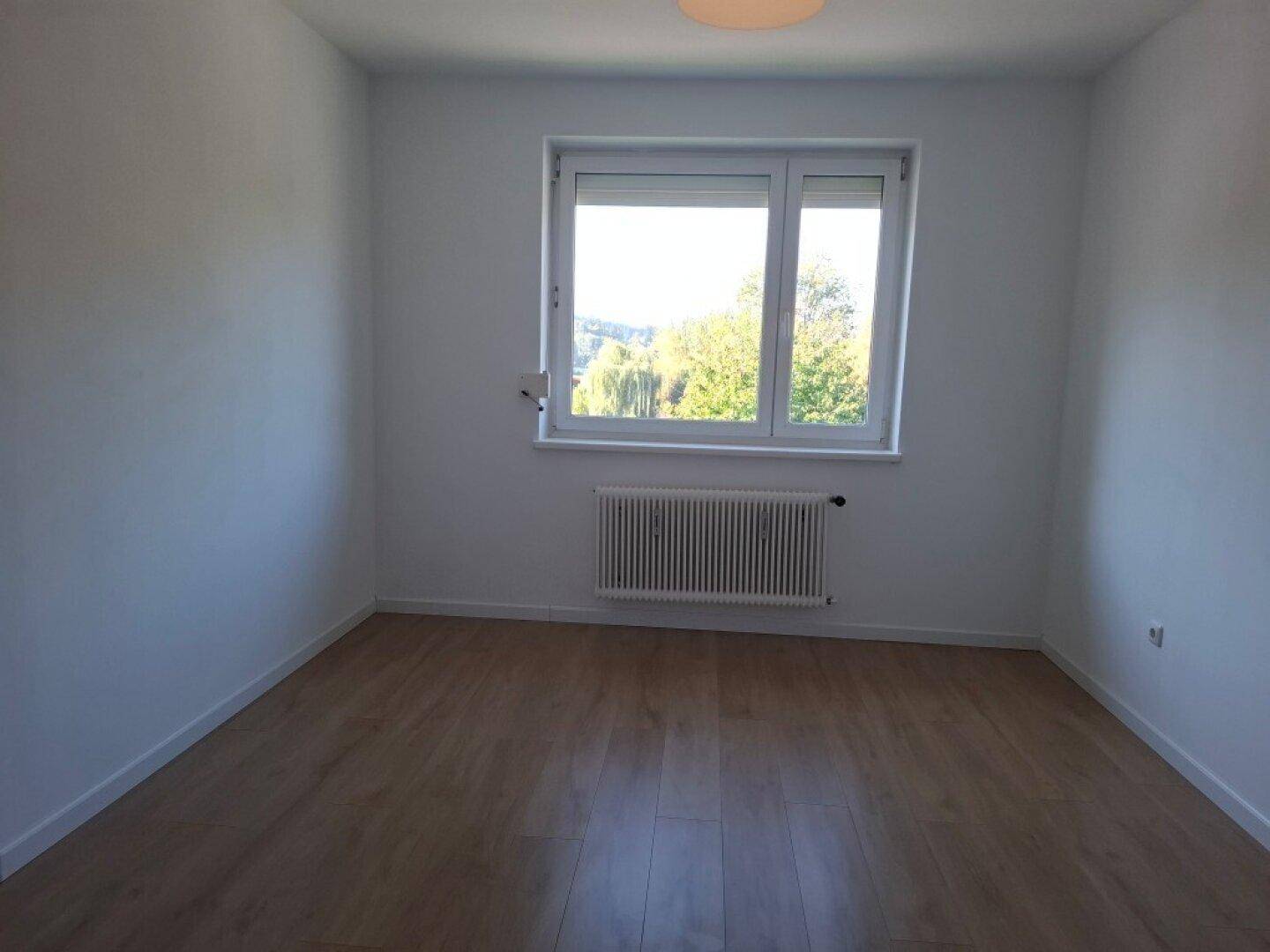 Leeres Zimmer mit Laminatboden, einem Fenster und einem Heizkörper, bereit zur individuellen Gestaltung.