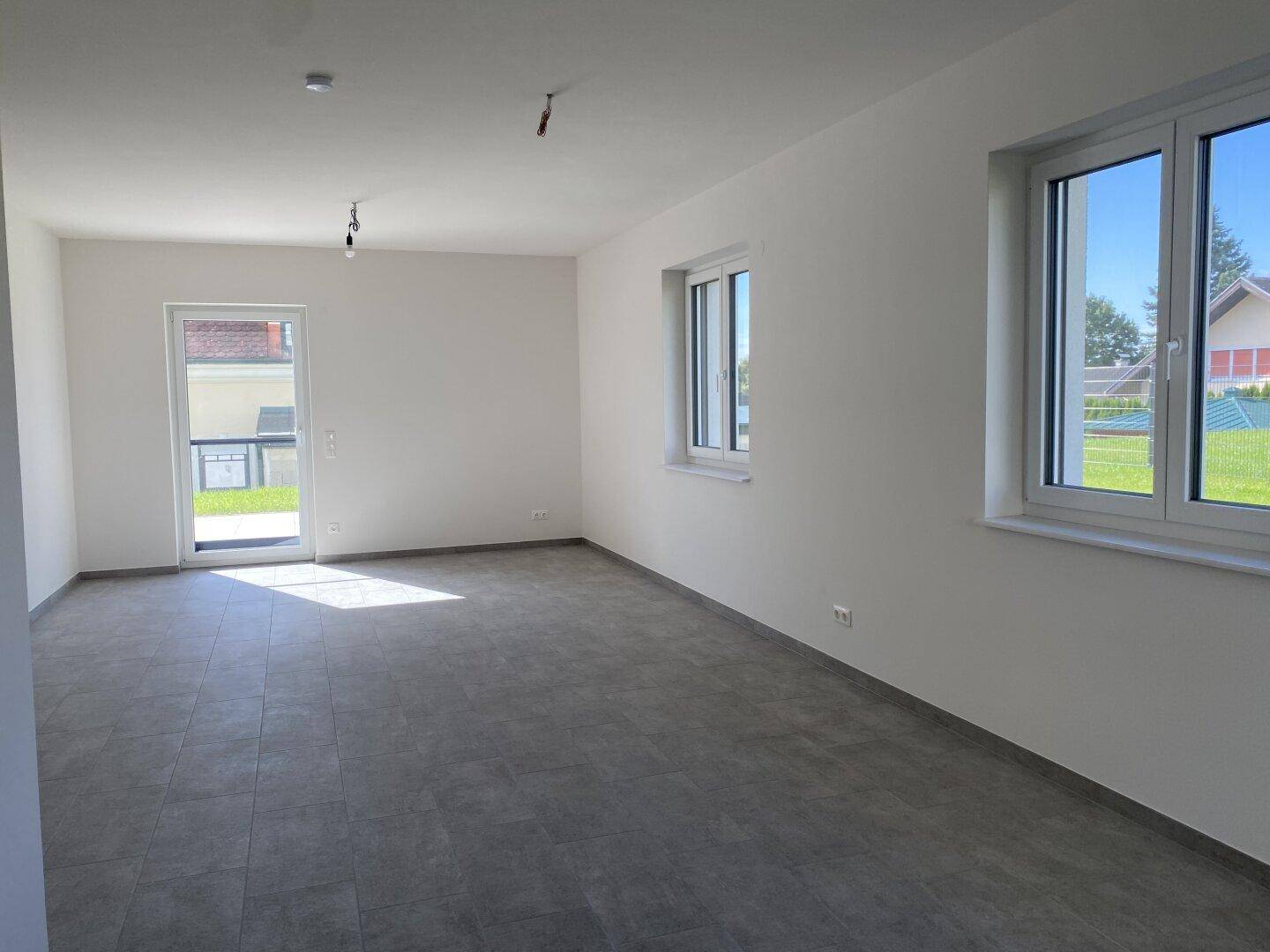 Heller, leerer Wohnraum mit grauen Fliesen, weißen Wänden und Zugang zur Terrasse durch eine Glastür.