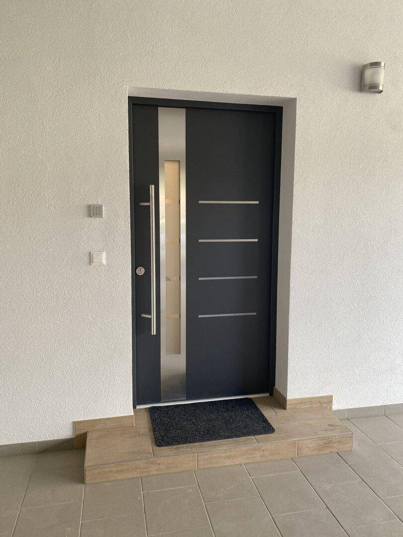Moderne Eingangstür in Anthrazit mit Glaseinsätzen und langem Edelstahlgriff, auf gefliestem Podest.