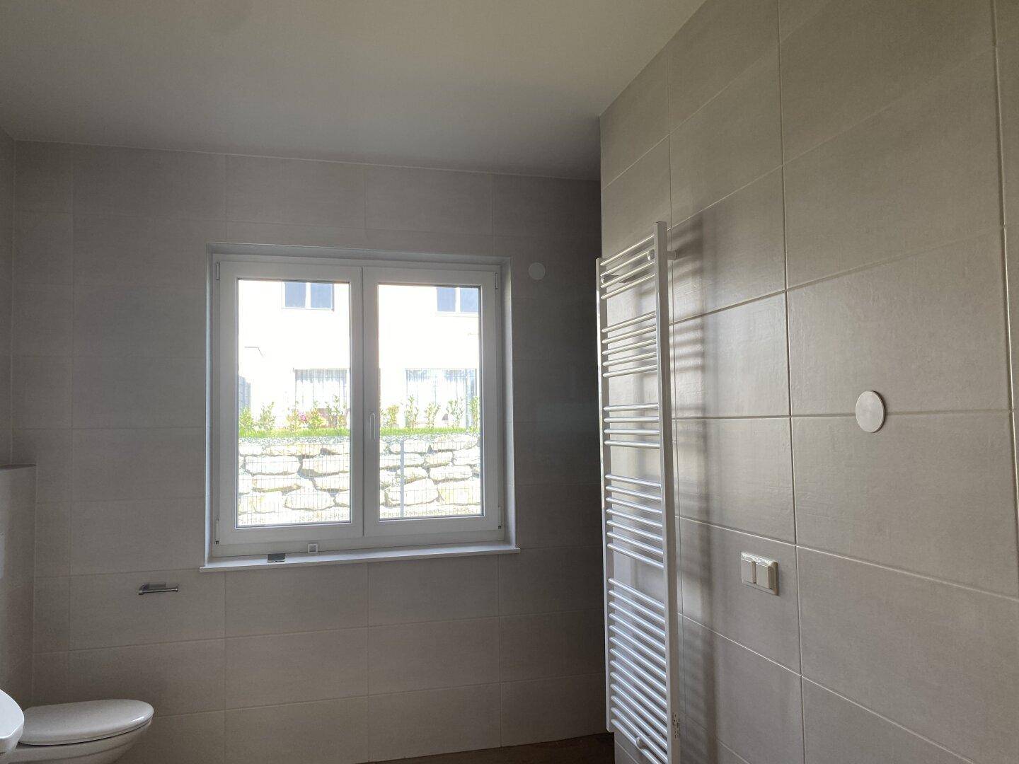 Helles Badezimmer mit Fenster, grauen Fliesen und einem praktischen Handtuchheizkörper an der Wand.