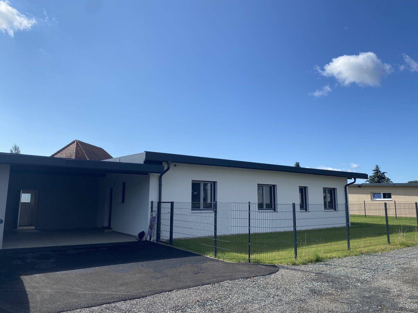 Seitenansicht des modernen Hauses mit integriertem Carport und umzäuntem Grundstück unter blauem Himmel.