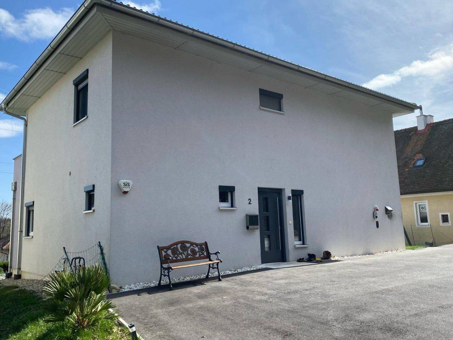 Moderne Hausfassade in Grau mit dunkler Eingangstür und kleinen Fenstern, umgeben von einer gepflegten Auffahrt.