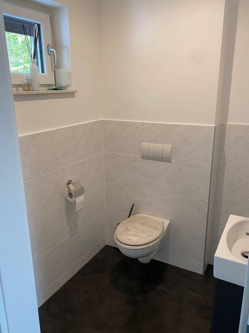 Helles Gäste-WC mit wandhängender Toilette, kleinem Waschbecken und einem Fenster für Tageslicht.