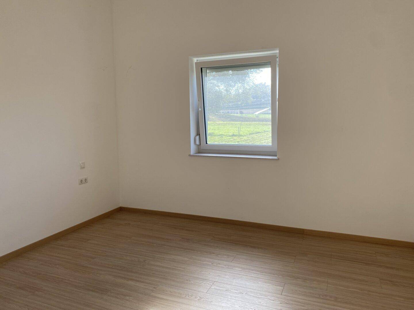 Leeres Zimmer mit hellem Laminatboden und einem Fenster, das viel Tageslicht hereinlässt.