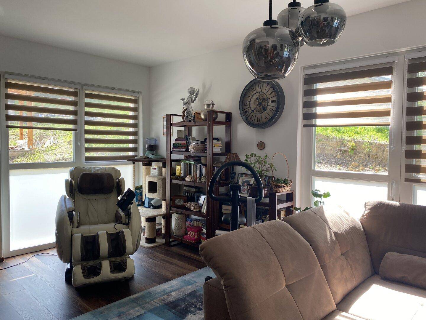 Wohnzimmer mit Massagesessel, Bücherregal und großen Fenstern mit Blick ins Grüne.