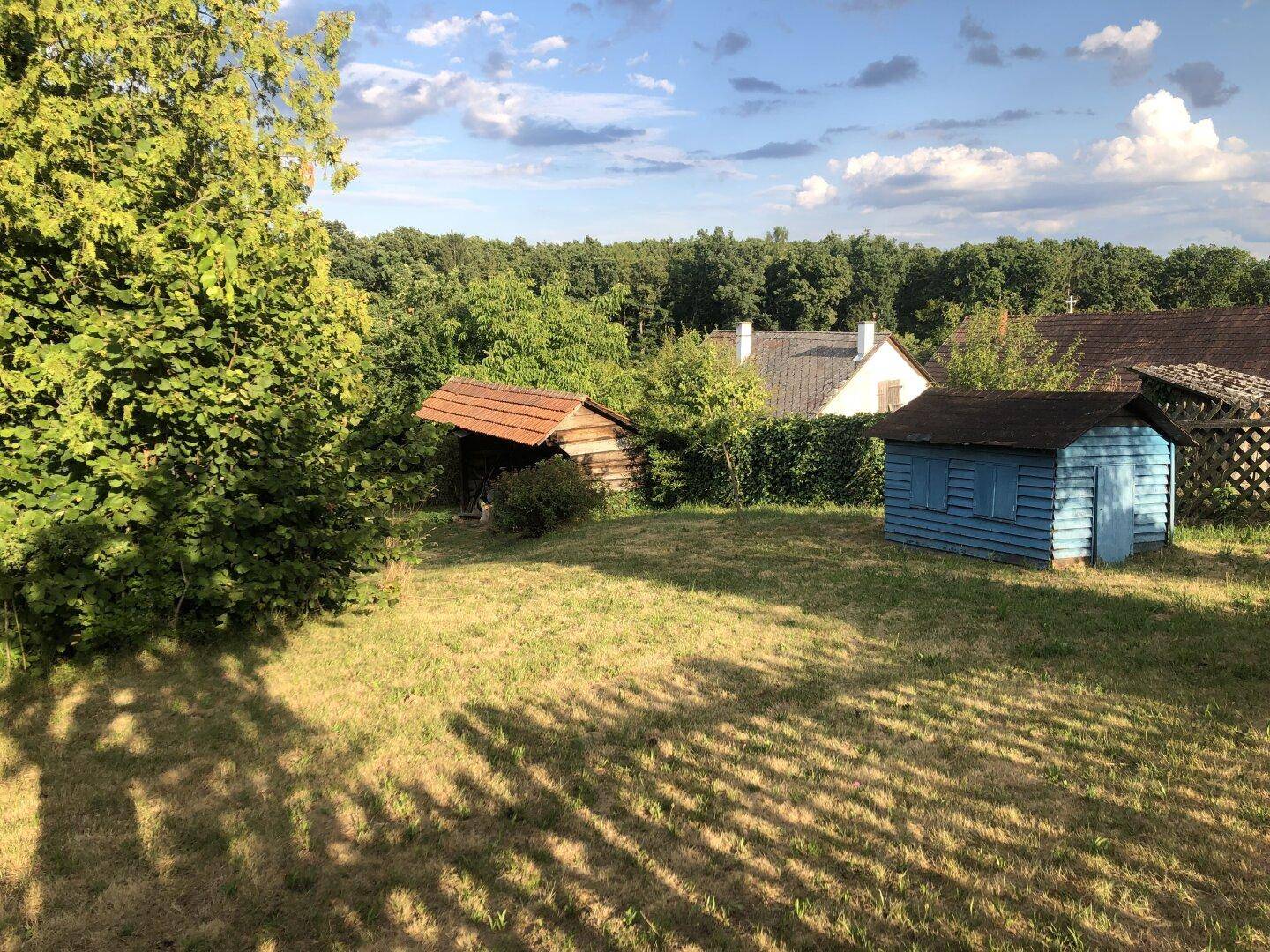 Großer Gartenbereich mit zwei kleinen Schuppen und Blick auf bewaldete Hügel.