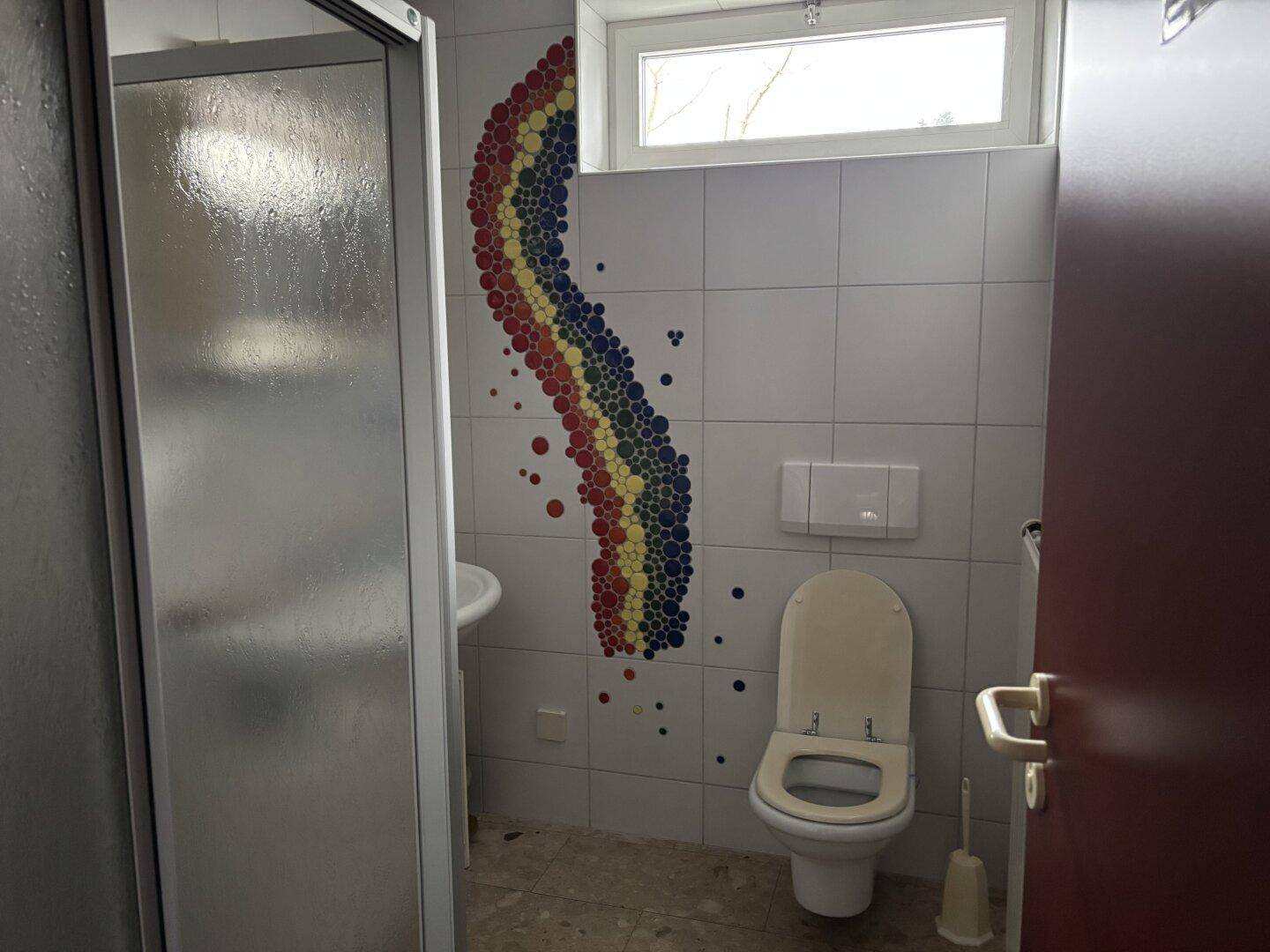 Badezimmer mit Dusche, Toilette und farbenfroher Mosaikdekoration an der Wand.