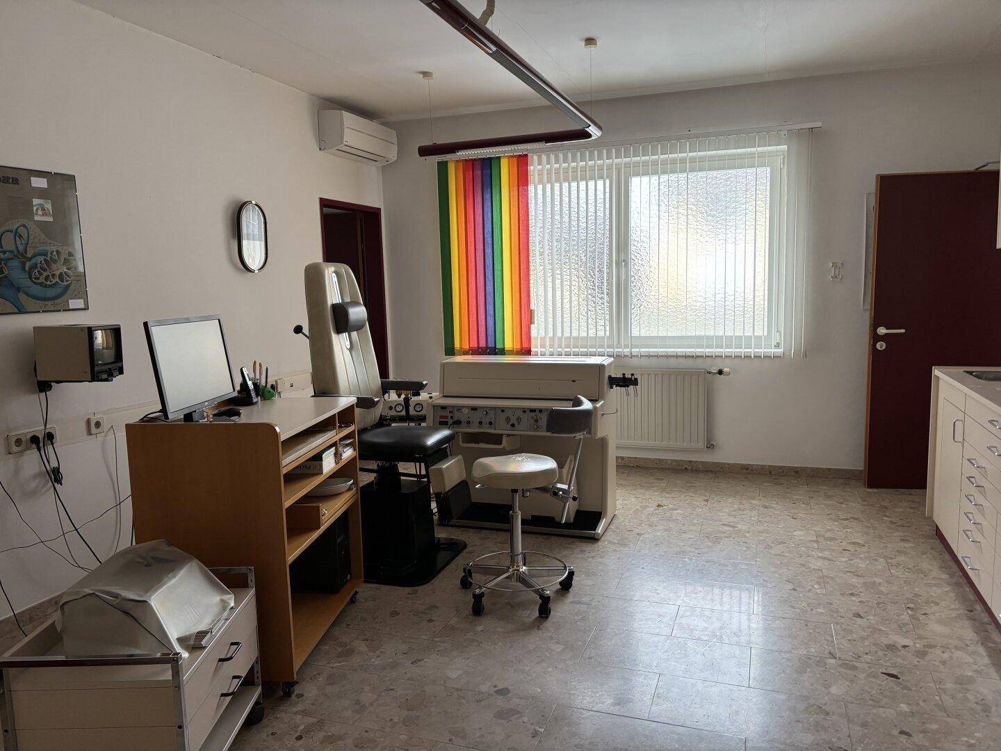 Behandlungszimmer mit medizinischen Geräten, Schreibtisch und Fenster mit Jalousien.