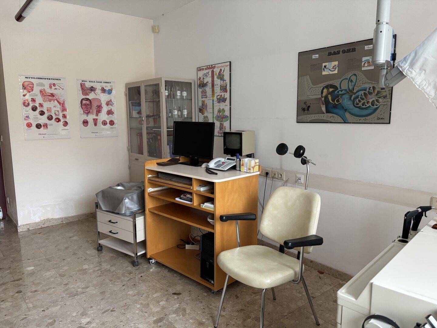 Behandlungszimmer mit Schreibtisch, Stuhl, medizinischen Diagrammen und Schränken.