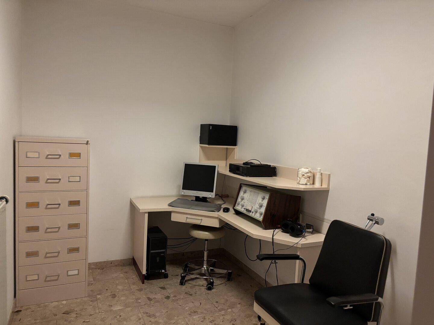 Behandlungszimmer mit Schreibtisch, Computer, medizinischen Geräten und Aktenschrank.