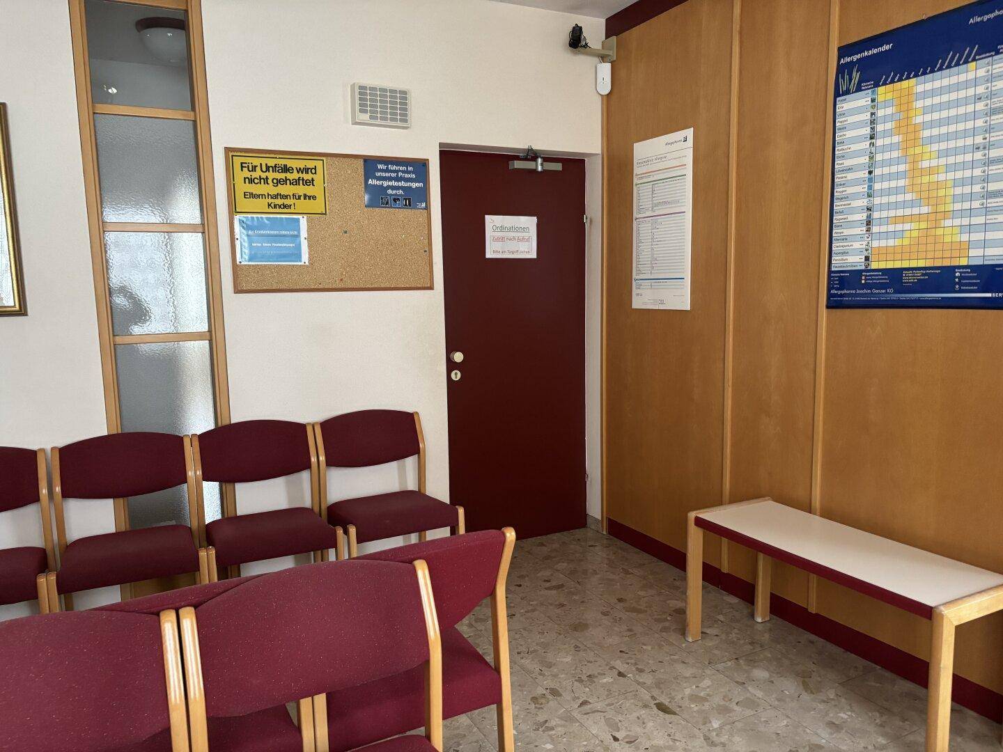 Wartezimmer mit Sitzbänken, einer Pinnwand und einer Tür, die zu weiteren Räumen führt.