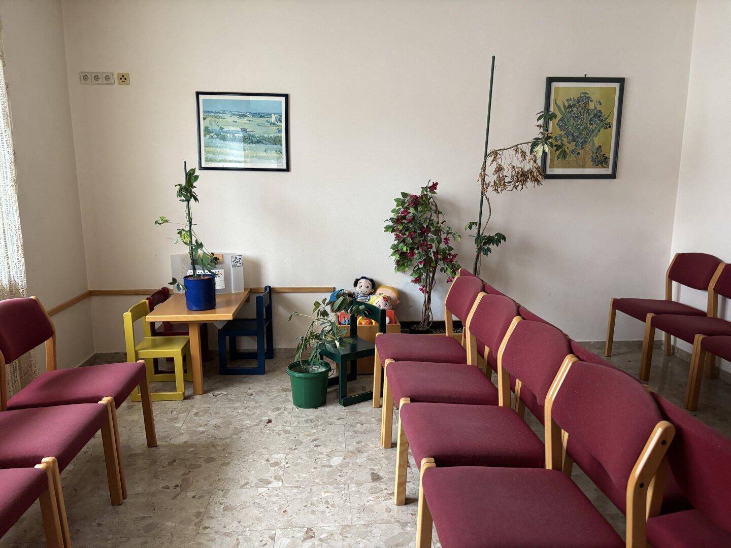 Wartezimmer mit Reihen von Stühlen, einem Kinderspielbereich und gefliestem Boden.