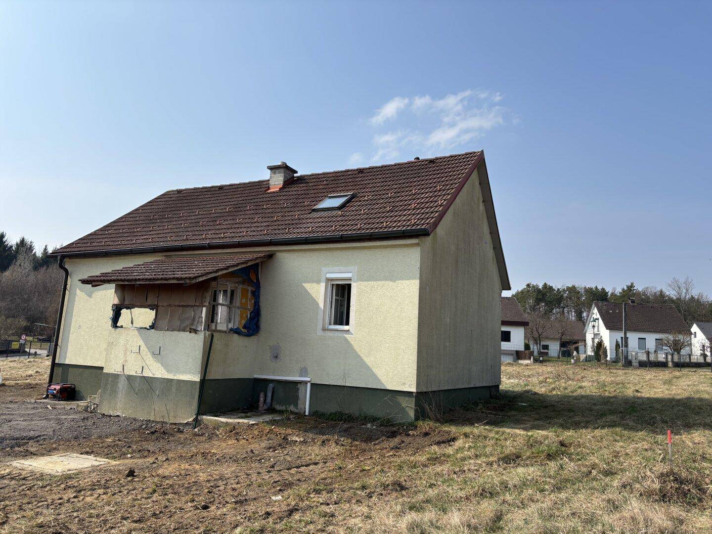 Seitenansicht eines Hauses mit beiger Fassade und rotem Ziegeldach, umgeben von trockenem Gras.