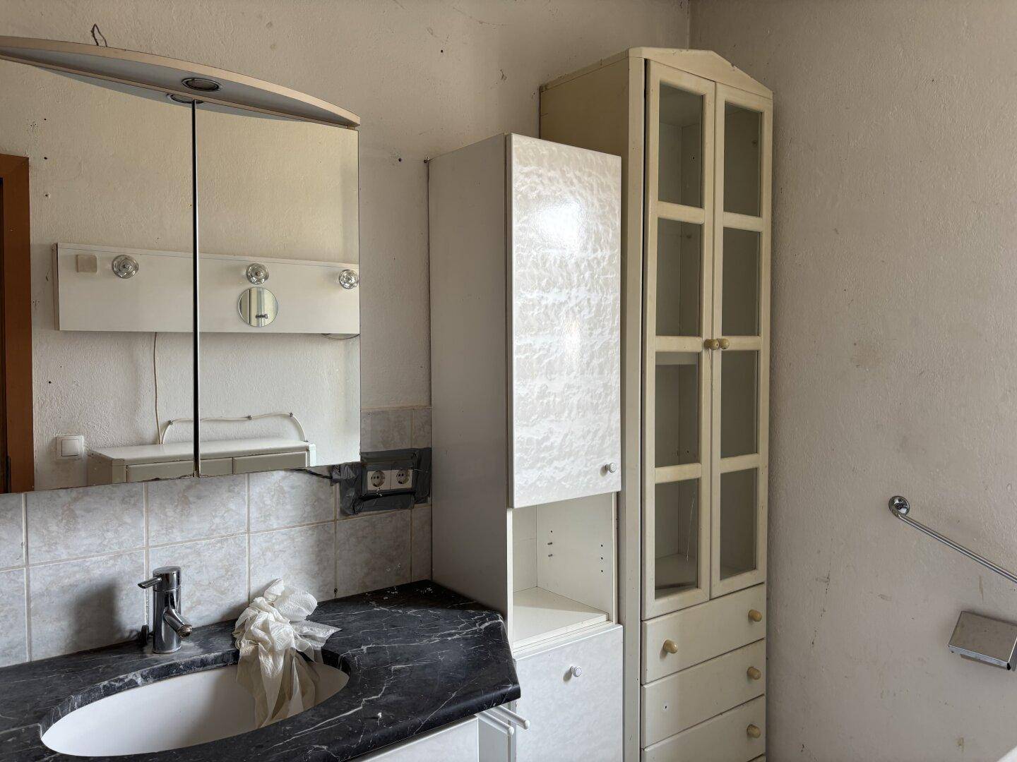 Badezimmer mit Waschbecken, Spiegelschrank und älteren Einbauschränken, renovierungsbedürftig.