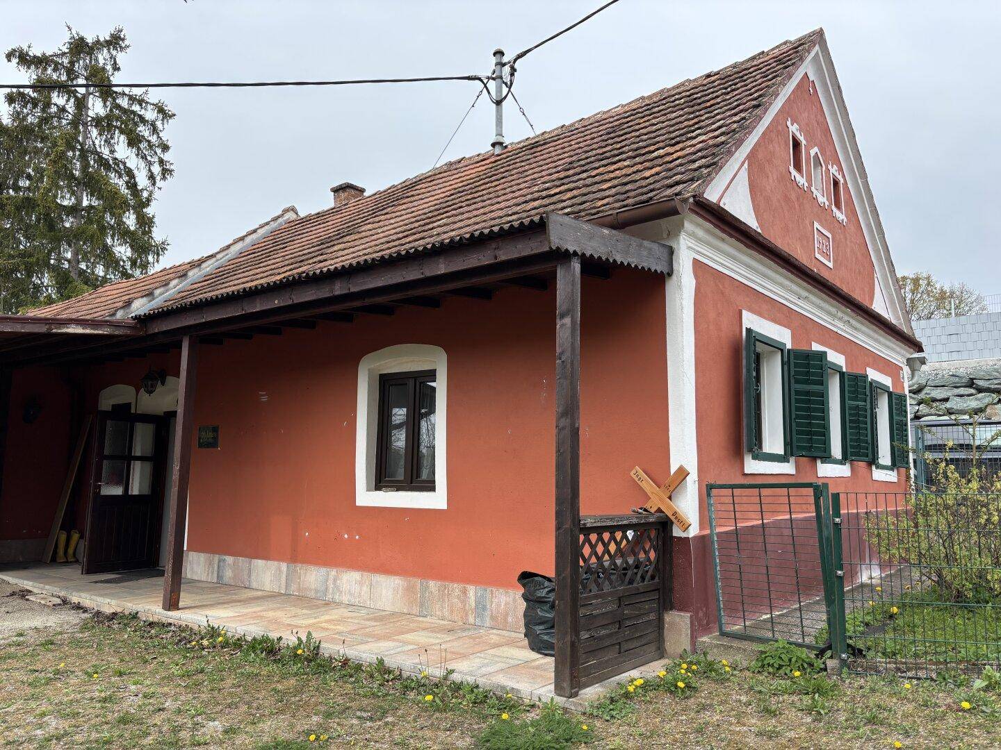 Seitenansicht des Hauses mit roter Fassade, grünen Fensterläden und überdachtem Eingang.