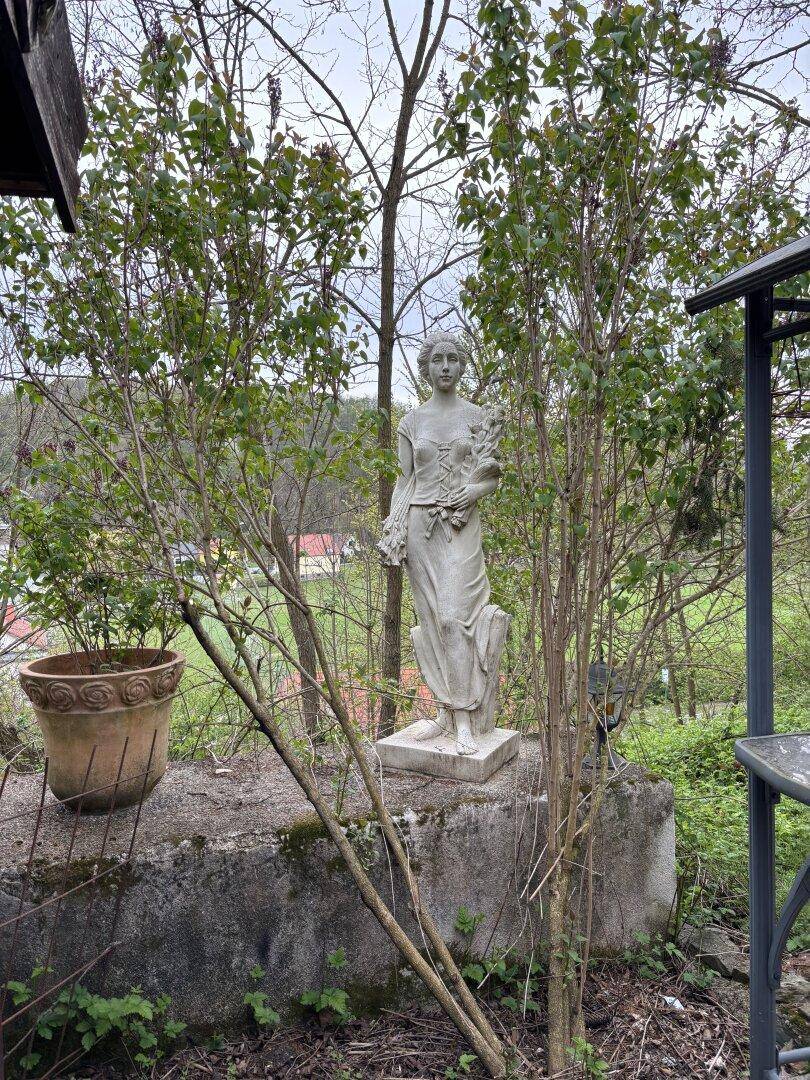 Gartenbereich mit klassischer Statue und üppiger Vegetation im Hintergrund.