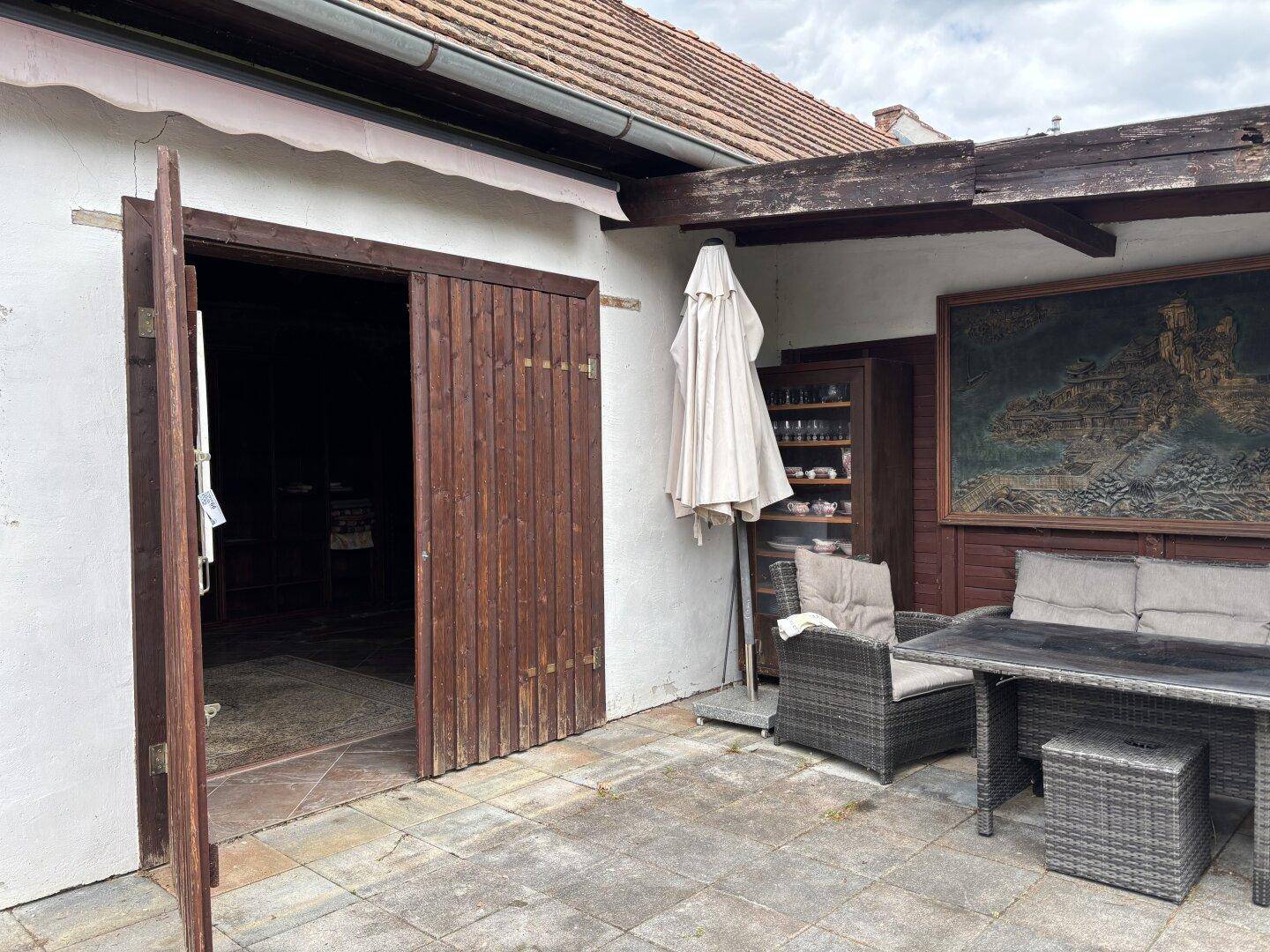 Gepflasterte Terrasse mit geöffneten Holztüren, Rattanmöbeln und einem großen Wandbild.