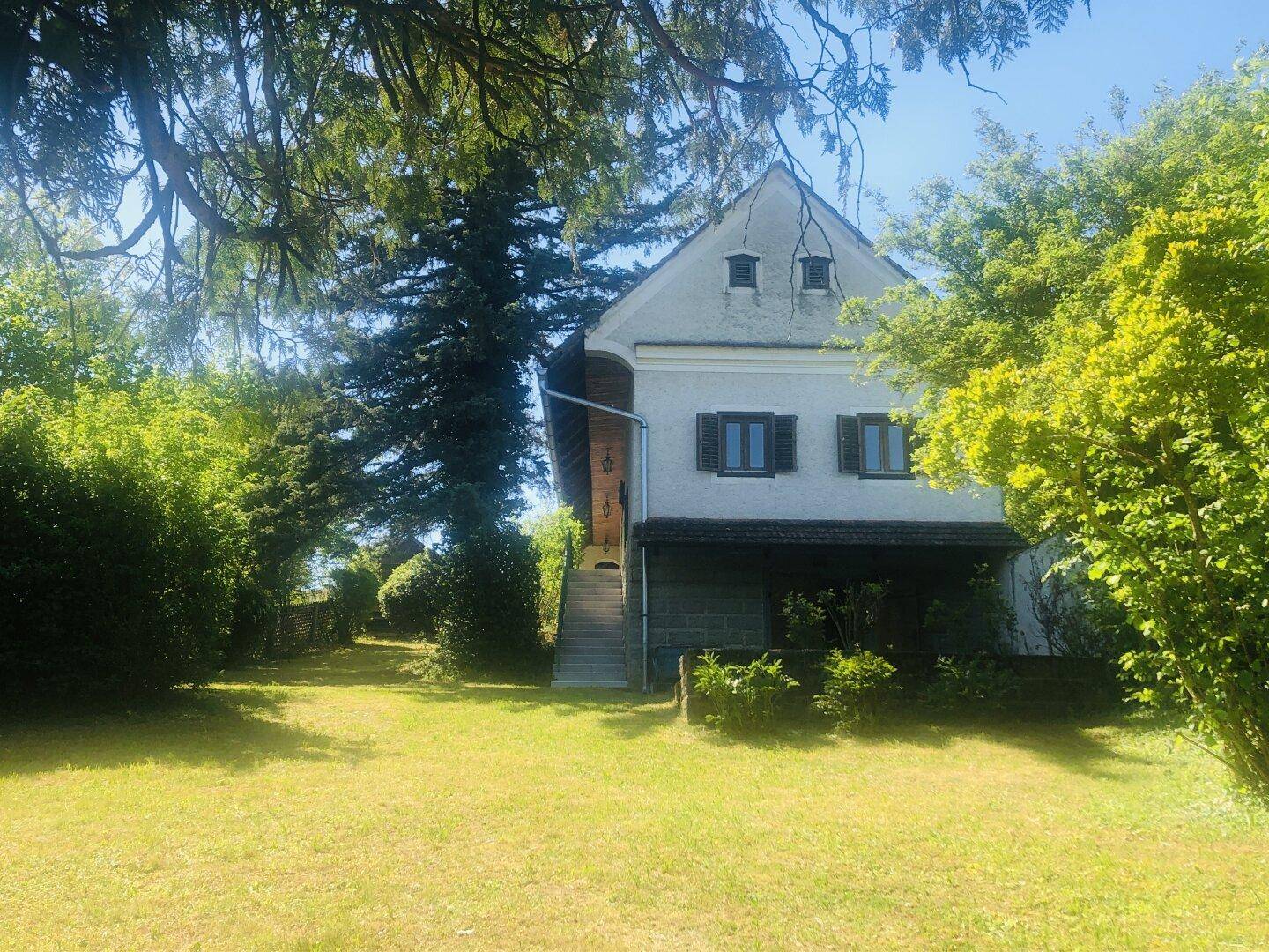 Traditionelles Landhaus mit großem Garten und altem Baumbestand unter blauem Himmel.