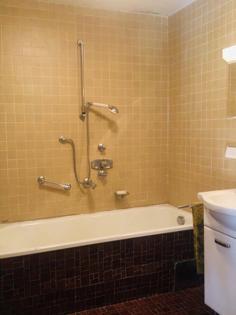 Badezimmer im 80er-Jahre-Stil mit beigefarbenen Fliesen, Badewanne und Duschvorrichtung.
