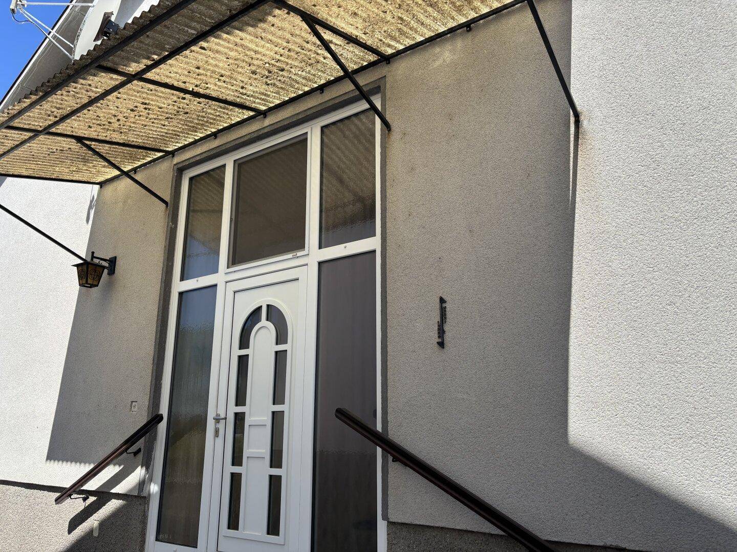 Eingangsbereich eines Hauses mit weißer Tür, Glaspaneelen und einem Vordach aus Wellblech.