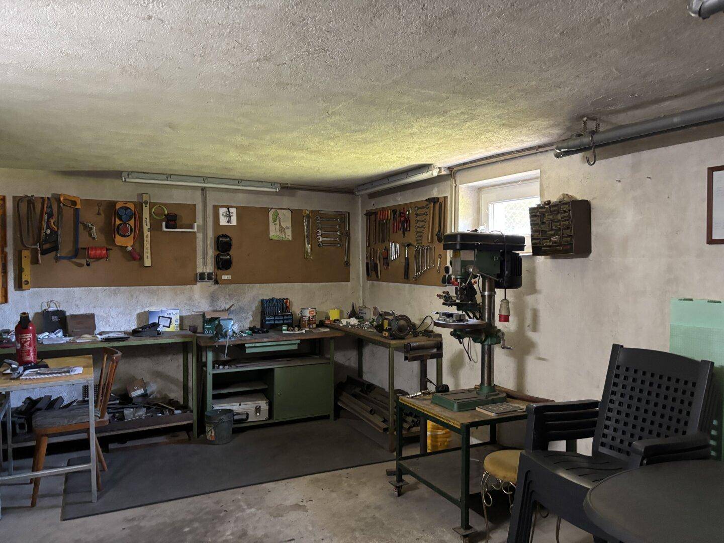 Werkstatt in einem Kellerraum mit Werkbänken, Werkzeugen und einer Bohrmaschine.