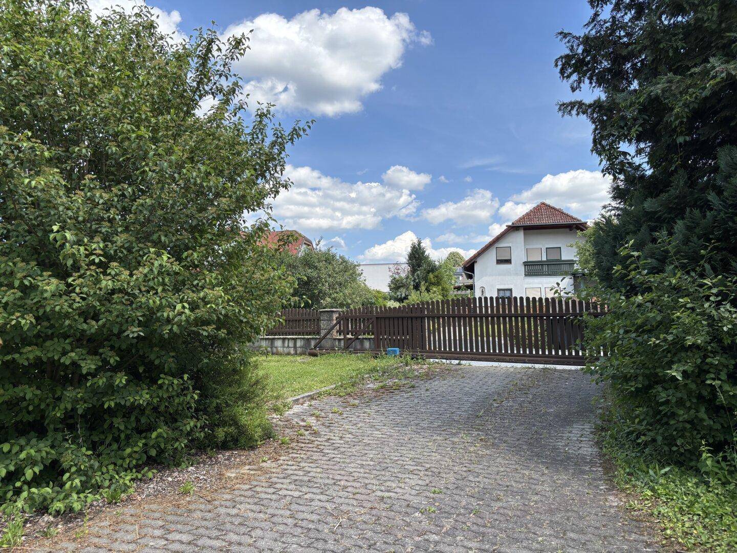 Gepflasterte Auffahrt mit Holzzaun und Bäumen, die zu einem Wohnhaus führt.