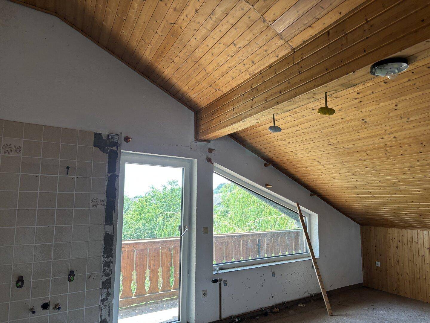 Dachgeschossraum mit Holzdachschrägen, Balkonzugang und vorbereiteten Küchenanschlüssen.