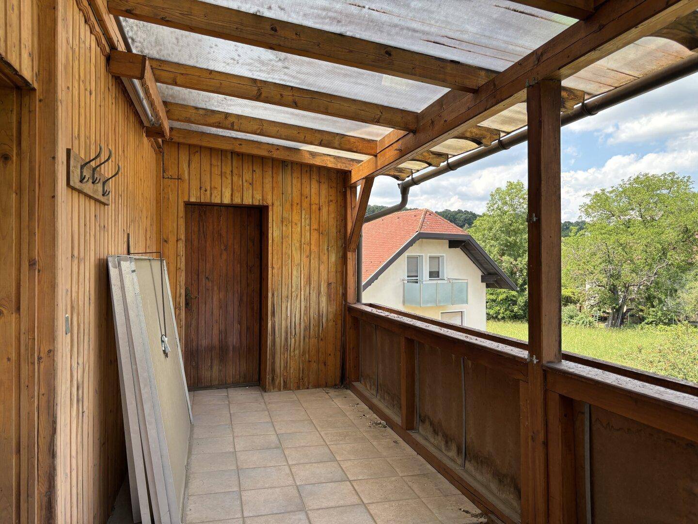 Überdachter Balkon mit Holzverkleidung, Fliesenboden und Blick ins Grüne.