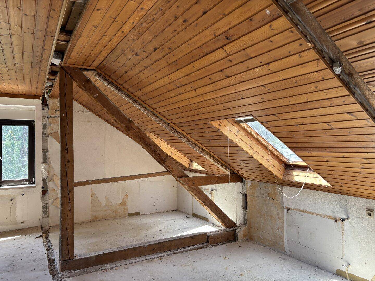 Dachgeschossraum im Rohbau mit sichtbaren Holzbalken, Holzdecke und Dachfenster.