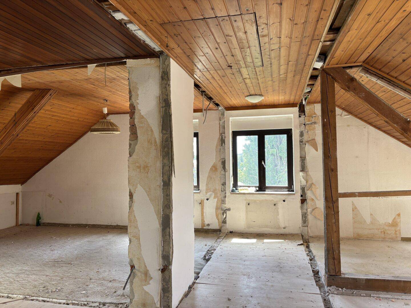 Großer Dachgeschossraum im Umbau mit Holzdecke, freigelegten Wänden und Fenstern mit Weitblick.