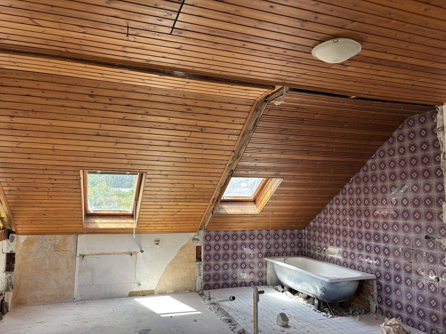 Badezimmer im Rohbau mit Holzdecke, zwei Dachfenstern und alter Badewanne.