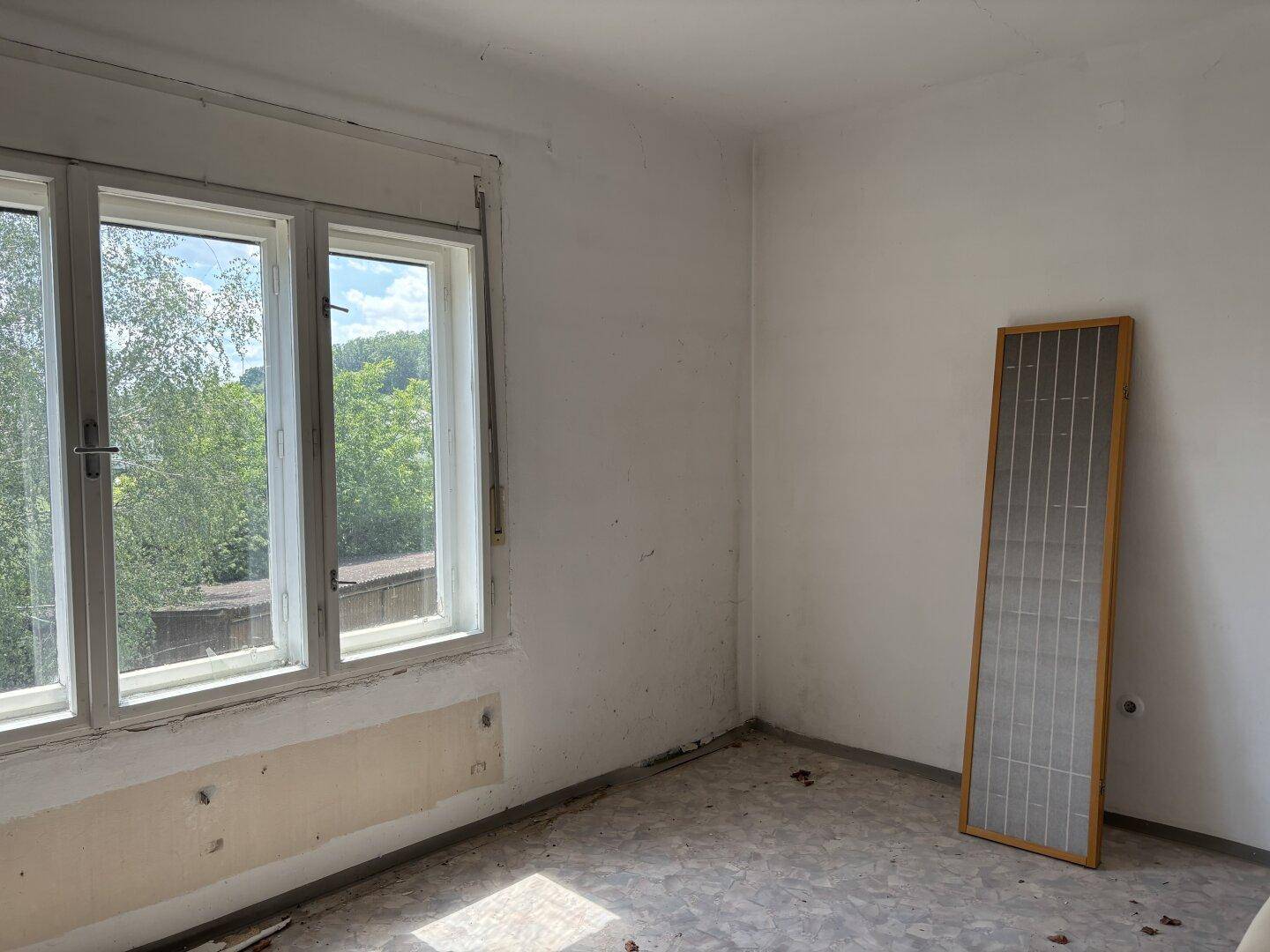 Leeres Zimmer mit weiß gestrichenen Wänden, einem Fenster und Blick ins Grüne.