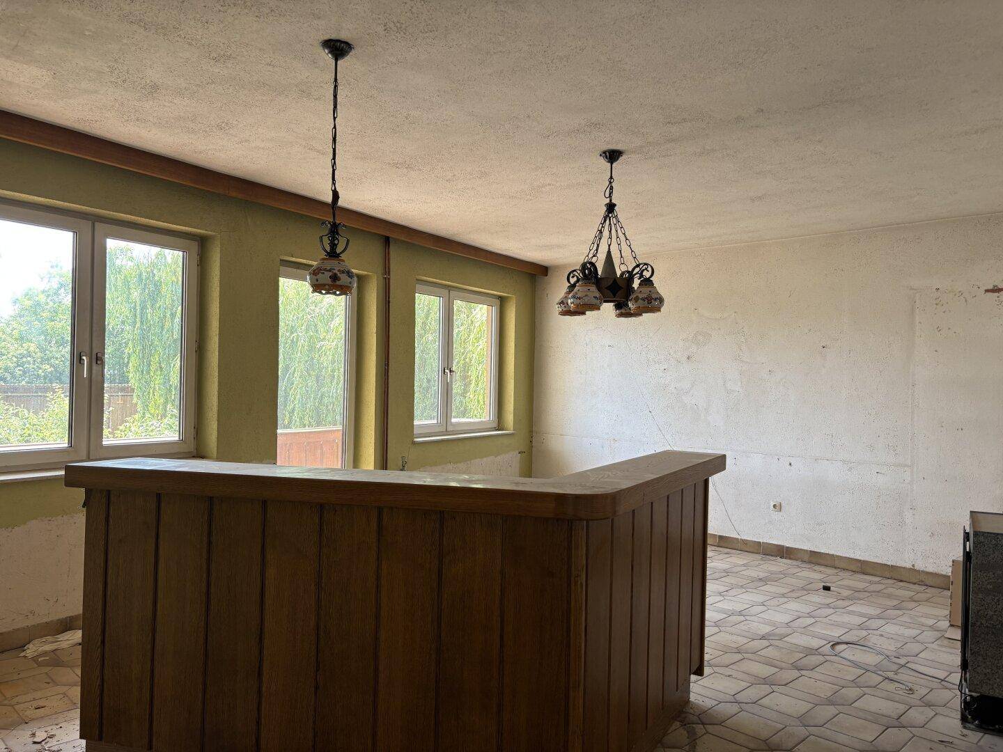 Großer Raum mit einer Holzbar, Fliesenboden und mehreren Fenstern mit Blick ins Grüne.