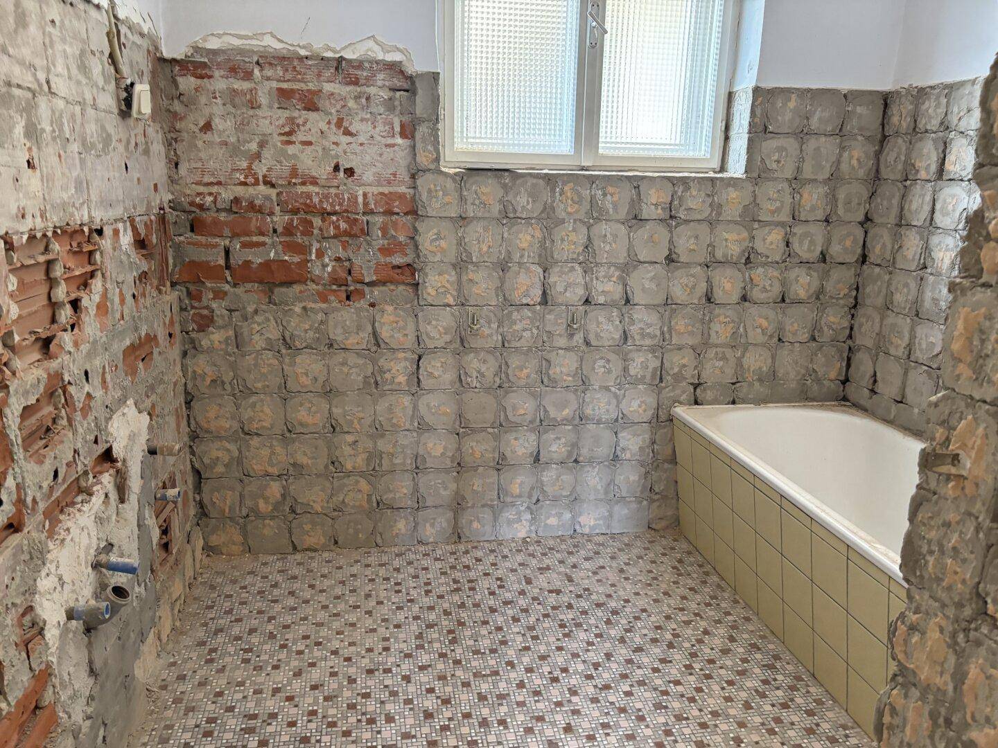 Badezimmer im Rohbau mit Badewanne, Mosaikfliesen und sichtbaren Ziegelwänden.