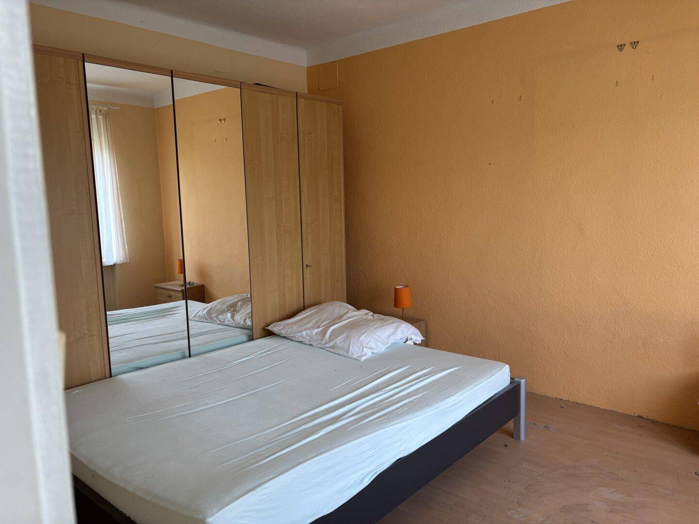Schlafzimmer mit Bett, Spiegelschrank und orangefarbenen Wänden, bereit zur Renovierung.