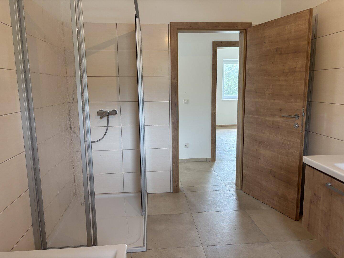 Helles Badezimmer mit beigen Fliesen, Duschkabine und Zugang zu weiteren Räumen.