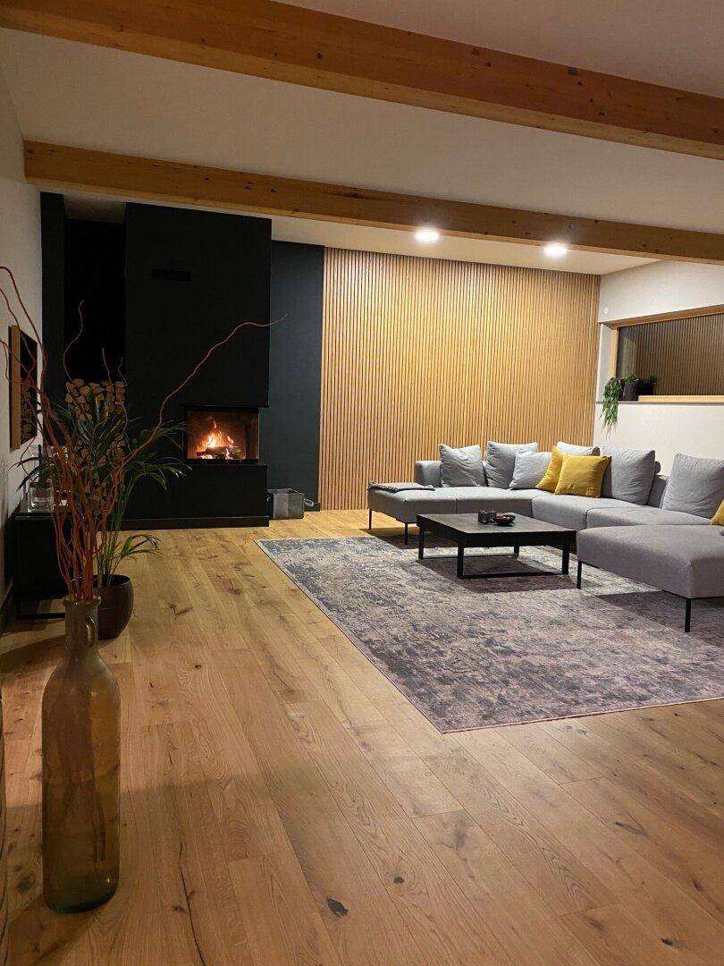 Gemütliches Wohnzimmer am Abend mit Kaminfeuer, Holzboden und sichtbaren Deckenbalken.