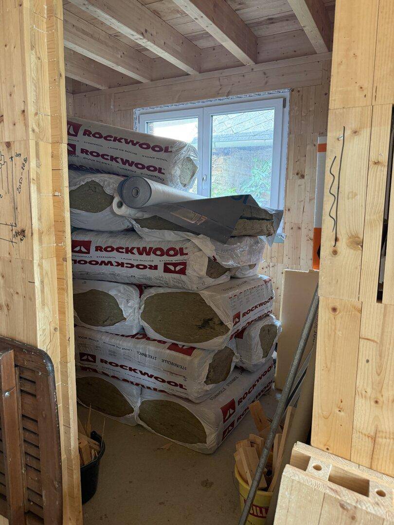 Abstellraum im Rohbau mit Holzwänden und gelagertem Dämmmaterial vor einem Fenster.