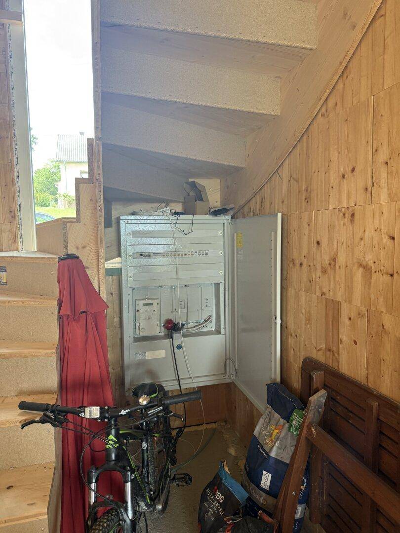 Unfertiger Flurbereich mit Holztreppe und offenem Elektroschaltkasten im Rohbauzustand.