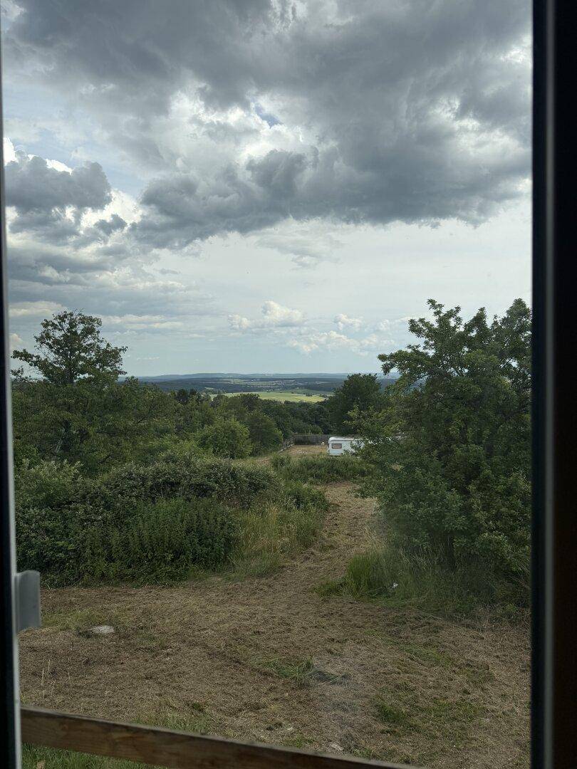 Blick aus dem Fenster auf eine grüne Landschaft mit Bäumen und einem Wohnwagen in der Ferne.