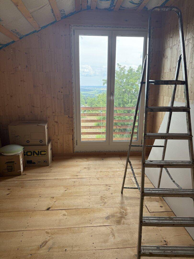 Geräumiges Dachzimmer mit Holzdielenboden und großem Fenster mit Panoramablick ins Grüne.
