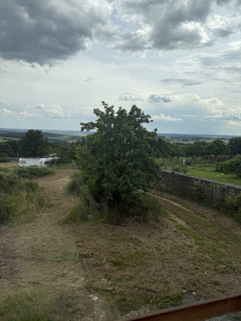Panoramablick über eine hügelige Landschaft mit Bäumen, einer Steinmauer und einem Wohnwagen.