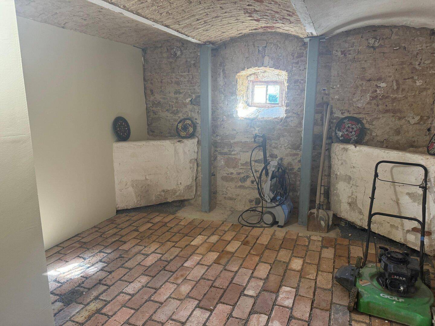Rustikaler Kellerraum mit Ziegelboden, unverputzten Steinwänden und einem kleinen Fenster.