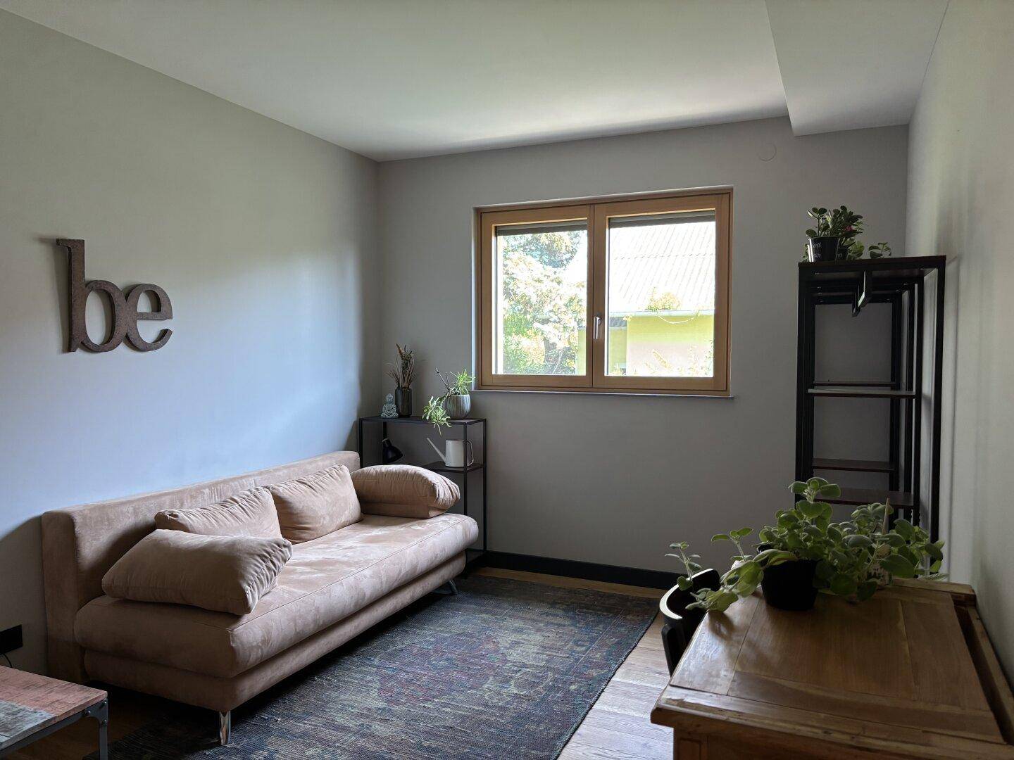Helles Zimmer mit Sofa, Fenster und einem Regal, ideal als Gäste- oder Arbeitszimmer.