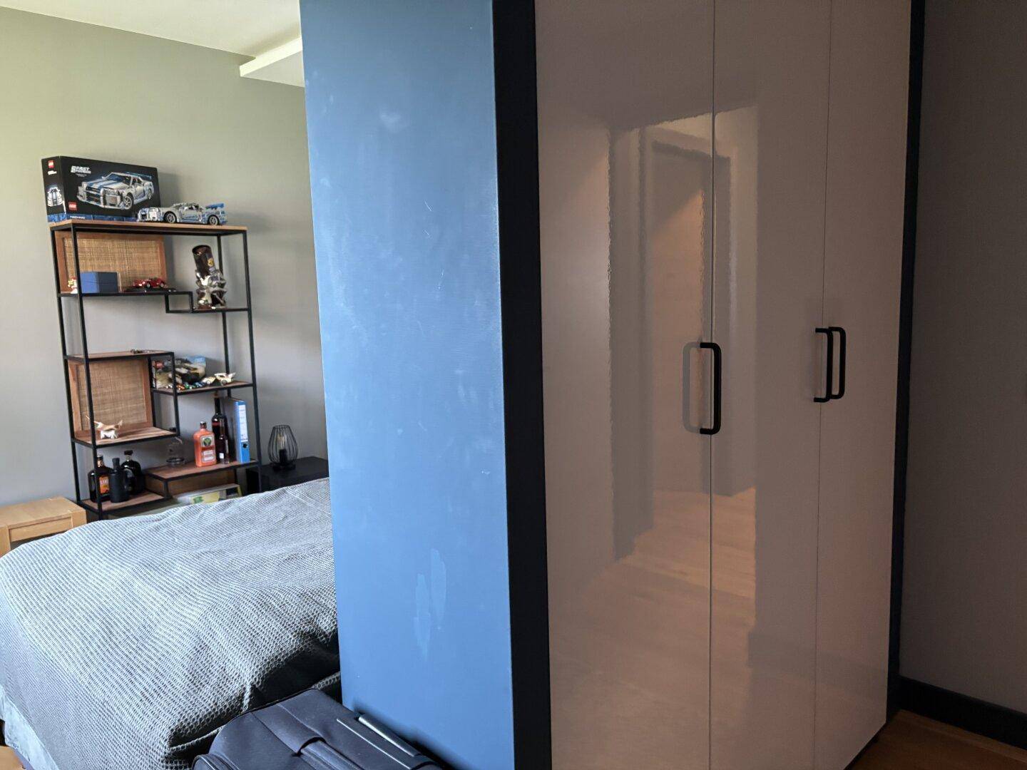 Schlafzimmer mit Bett, Regal und einem weißen Kleiderschrank, akzentuiert durch eine blaue Wand.