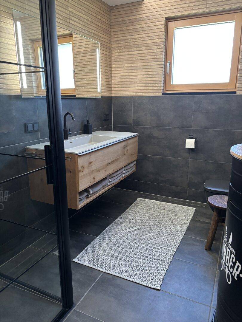 Modernes Badezimmer mit schwebendem Holzwaschtisch, dunklen Fliesen und einer begehbaren Dusche.