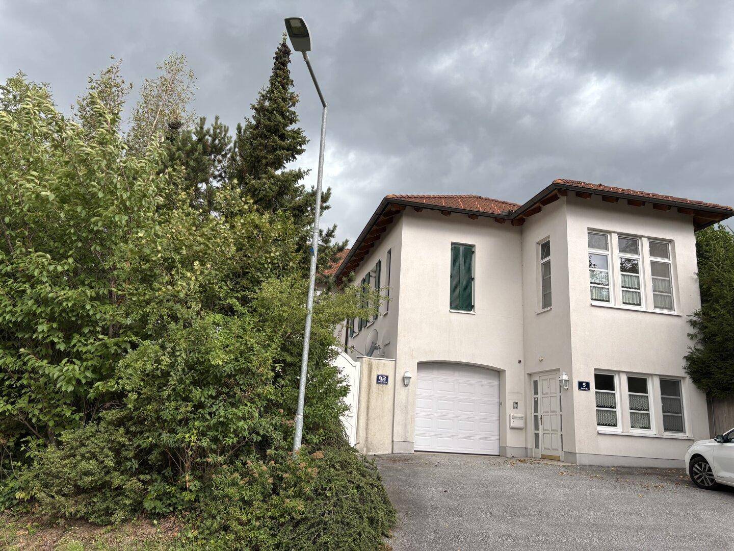 Gesamtansicht des weißen Hauses mit üppiger Begrünung, Zufahrt und einem Laternenpfahl.