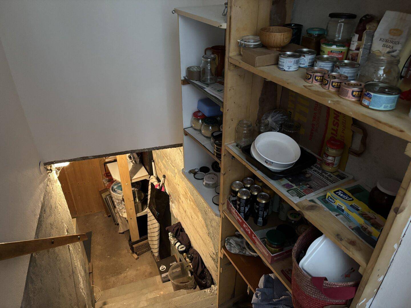 Blick in einen Kellerraum mit Holzregalen voller Vorräte und einer einfachen Treppe.