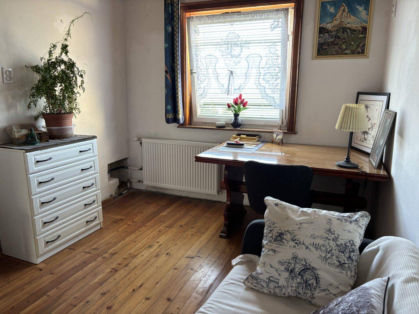 Helles Zimmer mit Holzboden, Schreibtisch am Fenster und einem kleinen Sofa, ideal zum Arbeiten.