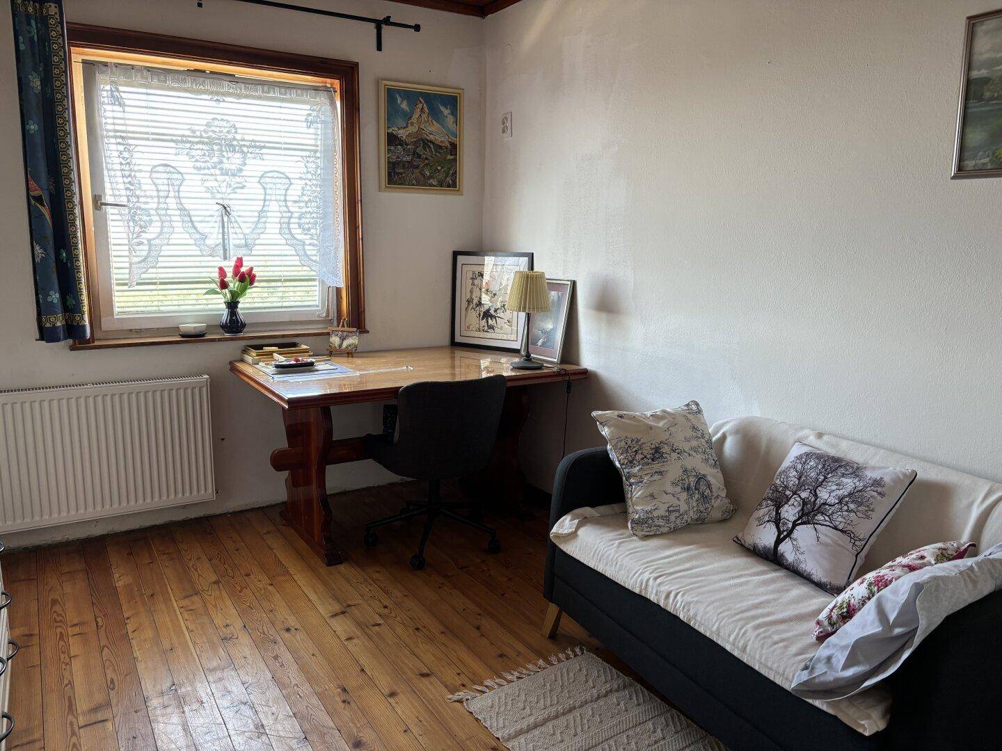 Arbeitszimmer mit Holzboden, Schreibtisch am Fenster und einem kleinen Sofa, gemütlich eingerichtet.